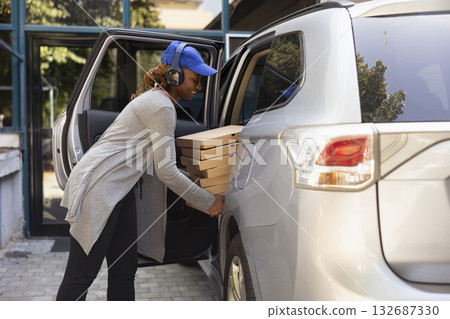 Black takeout worker delivering boxes of pizza at a location, driving the delivery car for a huge fast food order. Woman courier ensuring an express city service takeaway, drop off. Black takeout worker delivering boxes of pizza at a location, driving the delivery car for a huge fast food order. Woman courier ensuring an express city service takeaway, drop off. 132687330