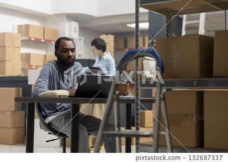 Black warehouse worker handling cargo and shipping logistics, coordinating inventory and hand packing merchandise, industrial warehouse shelves. Express delivery service at desk. Black warehouse worker handling cargo and shipping logistics, coordinating inventory and hand packing merchandise, industrial warehouse shelves. Express delivery service at desk. 132687337