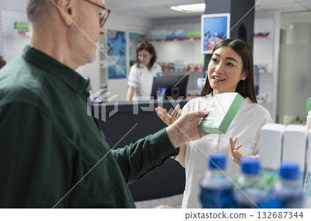 Asian pharmacist offering self care advice and vitamins to old person, recommending wellness supplements to help with mobility. Apothecary pharmacy visit with professional worker. Asian pharmacist offering self care advice and vitamins to old person, recommending wellness supplements to help with mobility. Apothecary pharmacy visit with professional worker. 132687344
