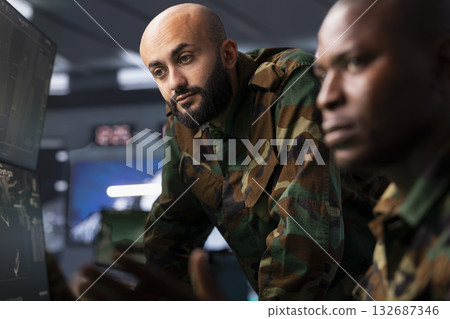 Team of military dispatchers looking at surveillance monitor, analyzing data to allocate resources to units based on mission needs. Agent and colleague examining strategic data on equipment displays Team of military dispatchers looking at surveillance monitor, analyzing data to allocate resources to units based on mission needs. Agent and colleague examining strategic data on equipment displays 132687346
