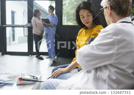 Doctor using blood pressure cuff to assess pulse and heartbeat during routine evaluation at modern clinic. Asian woman listens to advice while results are reviewed for hypertension control. Doctor using blood pressure cuff to assess pulse and heartbeat during routine evaluation at modern clinic. Asian woman listens to advice while results are reviewed for hypertension control. 132687351