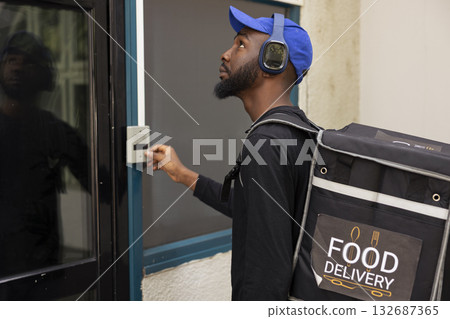 Black young employee arriving at the address for food delivery, ringing the door bell and intercom to deliver the meal order. Courier carrying the thermal backpack, city service. 132687365