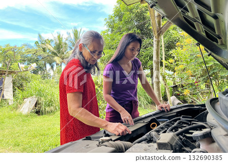 An elderly and young Asian woman looks under the hood of a broken down car to find a problem with the engine 132690385