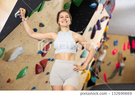 Positive slender girl in sportswear is standing near climbing wall in gym 132690669