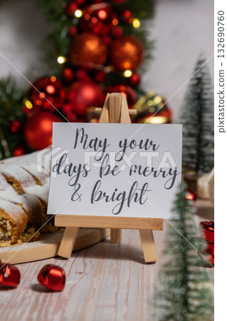 Greeting card with message MAY YOUR DAYS BE MERRY AND BRIGHT congratulations. Christmas Stollen in Christmas lights and decoration. Traditional sweet fruit loaf with icing sugar festive pastry dessert 132690760