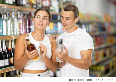 Man with wife near display of alcohol drinks in hypermarket doubt choice. Man with wife near display of alcohol drinks in hypermarket doubt choice. 132690872