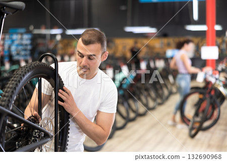 Young male customer choosing bike and checking tire on bicycle wheel in sport store 132690968