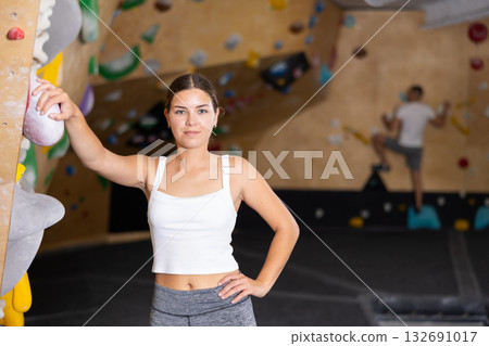 Positive slender girl in sportswear is standing near climbing wall in gym Positive slender girl in sportswear is standing near climbing wall in gym 132691017