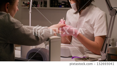 Manicure treatment. Spa therapy. Beautician female master hands in pink protective gloves mask using wooden stick to push back clients cuticle doing professional nail procedure. Manicure treatment. Spa therapy. Beautician female master hands in pink protective gloves mask using wooden stick to push back clients cuticle doing professional nail procedure. 132693041