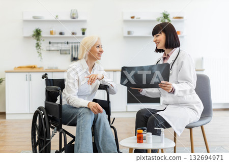 A female doctor shows an X-ray to an elderly patient in a wheelchair, discussing the results in a bright room. The patient smiles, indicating a positive interaction. 132694971