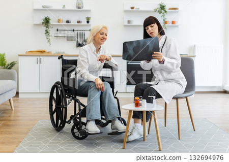 A female doctor shows an X-ray to an elderly woman in a wheelchair, discussing the results in a home setting. The patient is smiling while the doctor explains. 132694976