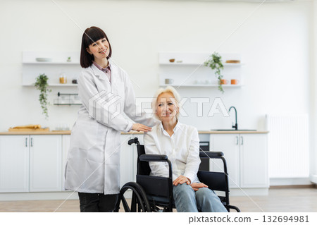 A young woman in a white coat stands beside an older woman in a wheelchair, both smiling. They are indoors in a bright, clean kitchen setting. A young woman in a white coat stands beside an older woman in a wheelchair, both smiling. They are indoors in a bright, clean kitchen setting. 132694981