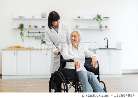 A smiling caregiver pushes an elderly woman in a wheelchair indoors, likely in a kitchen setting, offering support and care. A smiling caregiver pushes an elderly woman in a wheelchair indoors, likely in a kitchen setting, offering support and care. 132694984