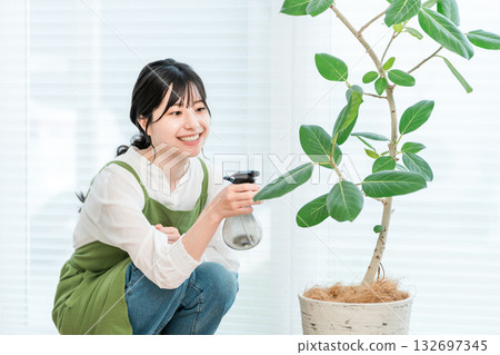 A young Asian woman wearing an apron spraying water on a houseplant 132697345
