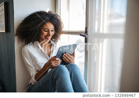 Portrait of young woman reading ebook, sitting on window sill. Portrait of young woman reading ebook, sitting on window sill. 132698467