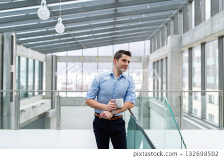 Young businessman standing indoors in an office, holding coffee cup. Young businessman standing indoors in an office, holding coffee cup. 132698502