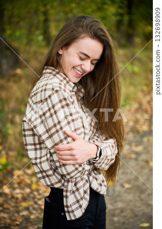 A young woman with long hair smiles warmly while standing in a colorful autumn forest, wearing a cozy plaid shirt and surrounded by falling leaves. 132698909
