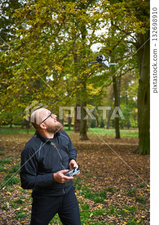 A man, deeply focused, controls a flying drone amidst vibrant autumn leaves in a lush park. The scene captures the joy of technology blending with nature's beauty. A man, deeply focused, controls a flying drone amidst vibrant autumn leaves in a lush park. The scene captures the joy of technology blending with nature's beauty. 132698910