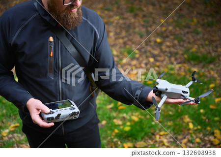 A man holds a drone in one hand and a controller in the other, ready to take off in a vibrant park filled with fallen leaves. 132698938