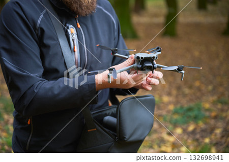 In a serene forest setting during autumn, a man carefully holds a drone, preparing for flight amidst fallen leaves and tall trees. 132698941