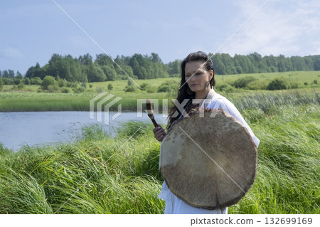 Female shaman in the white dress drumming outdoors 132699169