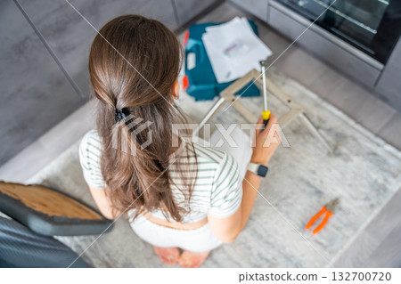 Young woman seen from behind assembling a chair with screwdrivers on the floor at home. Concept of modern lifestyle and practical skills in everyday life. Young woman seen from behind assembling a chair with screwdrivers on the floor at home. Concept of modern lifestyle and practical skills in everyday life. 132700720