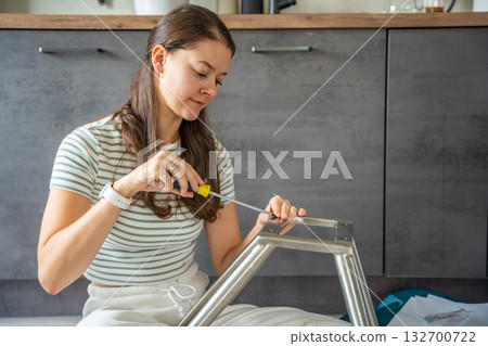 Smiling young woman assembling a chair with a screwdriver at home. Feminist concept celebrating female independence, confidence, and joy in do it yourself work. Smiling young woman assembling a chair with a screwdriver at home. Feminist concept celebrating female independence, confidence, and joy in do it yourself work. 132700722