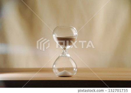 A simple hourglass on a wooden table with a beige backdrop, symbolizing time flow and minimalistic living. Represents timeless themes in a creative, uncluttered presentation for stylish concepts. 132701735