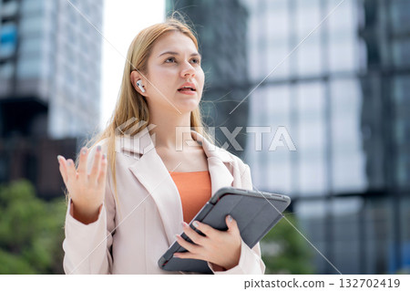 Professional young caucasian businesswoman talking using wireless earbud while holding tablet. 132702419