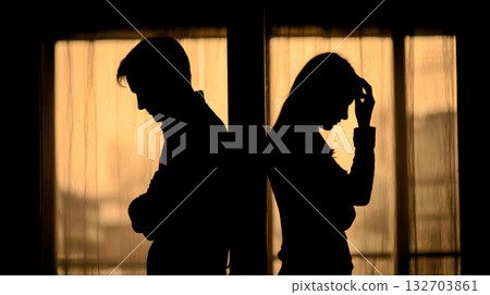 Sad couple struggles with emotions while standing back to back near a window at sunset Sad couple struggles with emotions while standing back to back near a window at sunset 132703861