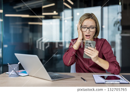 Young businesswoman with glasses feeling stressed and shocked while looking at her smartphone, reading bad news or having a problem at work in a modern office 132705765