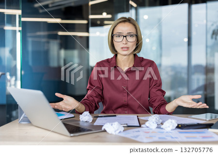 Businesswoman at her desk surrounded by crumpled papers and laptop, gesturing in frustration and confusion as she struggles with a stressful deadline and workplace pressure 132705776