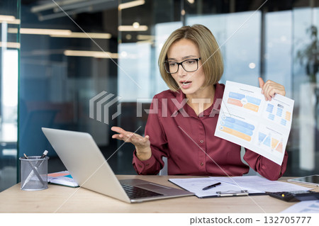 Businesswoman sitting at desk in modern office, having a virtual meeting on laptop, holding up financial charts and reports, and discussing data with a serious expression Businesswoman sitting at desk in modern office, having a virtual meeting on laptop, holding up financial charts and reports, and discussing data with a serious expression 132705777