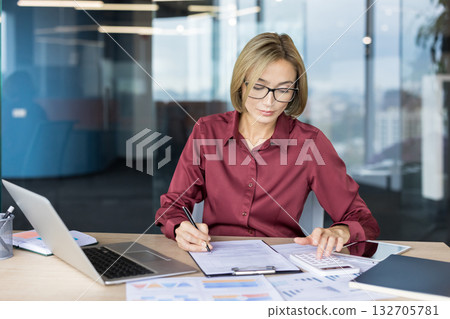 Woman accountant at office desk using calculator and laptop to analyze financial reports, manage budgets and paperwork, focused on numbers, planning and business strategy 132705781