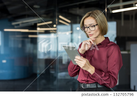 Businesswoman wearing glasses and a maroon shirt standing in a contemporary office, carefully reviewing data on a digital tablet with a focused and contemplative expression 132705796