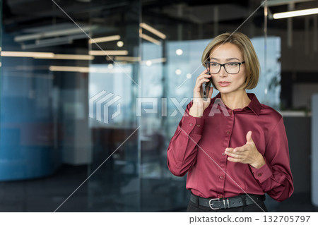 Professional businesswoman wearing glasses and a red shirt is having a phone conversation, actively gesturing and communicating in a modern corporate office environment 132705797