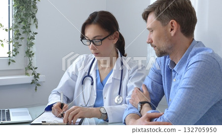 Female doctor consulting with patient in medical office using laptop and filling out paperwork to provide healthcare advice and support, discussing treatment options in a professional setting. Medical Female doctor consulting with patient in medical office using laptop and filling out paperwork to provide healthcare advice and support, discussing treatment options in a professional setting. Medical 132705950