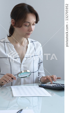 Accountant carefully examines financial documents, enlarges details with a magnifying glass, calculates performance indicators in a professional workplace. Audit and taxes in business. Accountant carefully examines financial documents, enlarges details with a magnifying glass, calculates performance indicators in a professional workplace. Audit and taxes in business. 132705952