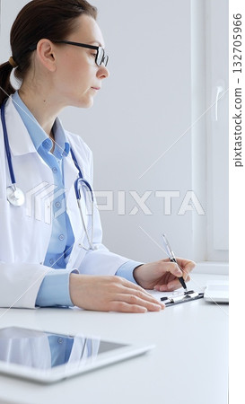 Adult female doctor completing healthcare paperwork, clipboard positioned near laptop and stethoscope in well lit clinical workspace by window. Medicine and health care Adult female doctor completing healthcare paperwork, clipboard positioned near laptop and stethoscope in well lit clinical workspace by window. Medicine and health care 132705966