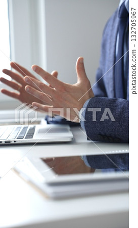 Businessman wearing suit and wristwatch sits at white desk explaining business strategy using hand gestures, with tablet and eyeglasses, vertical close up view. Business people concept Businessman wearing suit and wristwatch sits at white desk explaining business strategy using hand gestures, with tablet and eyeglasses, vertical close up view. Business people concept 132705967