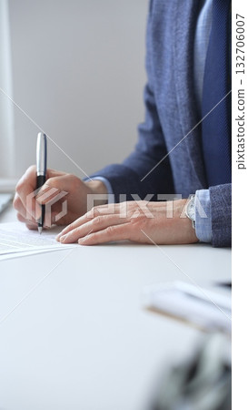 Close up of businessman's hands signing a contract with elegant pen, wearing a blue suit and a wristwatch, sitting at a white desk. Business people concept 132706007