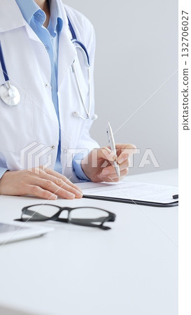 Unknown female doctor completing healthcare paperwork, clipboard positioned near laptop and stethoscope in well lit clinical workspace by window. Medicine and health care Unknown female doctor completing healthcare paperwork, clipboard positioned near laptop and stethoscope in well lit clinical workspace by window. Medicine and health care 132706027