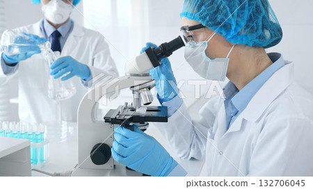 Masked female scientist examining specimen under microscope while laboratory assistant handles glassware in medical investigation at the background. Medicine, and science concept Masked female scientist examining specimen under microscope while laboratory assistant handles glassware in medical investigation at the background. Medicine, and science concept 132706045