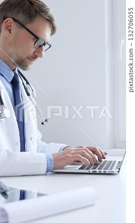 Adult doctor man is typing on a laptop, likely updating patient records or conducting research, showcasing the intersection of healthcare and technology in medical setting. Medicine concept 132706055