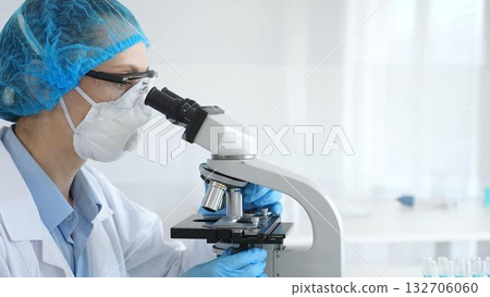Woman researcher in blue colored protective examining sample through microscope, conducting medical experiment inside sterile laboratory with glass vials surrounding workstation. Medicine and science 132706060