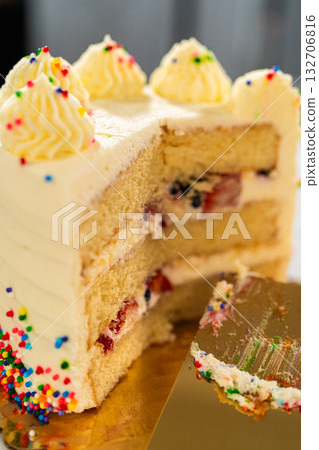A tall vanilla cake with piped frosting swirls and rainbow sprinkles stands partially sliced. A gold cake spatula rests in the slice. The background shows blurred dark tones for contrast. A tall vanilla cake with piped frosting swirls and rainbow sprinkles stands partially sliced. A gold cake spatula rests in the slice. The background shows blurred dark tones for contrast. 132706816
