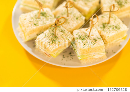 Delicate mini cucumber sandwiches made with fluffy white bread, a layer of herbed cream cheese, and thinly sliced cucumbers. Each square is topped with a sprinkle of fresh dill and secured with a 132706828