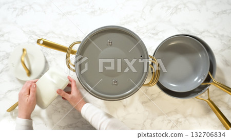 Hands reposition white ceramic cookware with gold handles on a marble countertop. The cookware pieces are neatly stacked and organized. 132706834