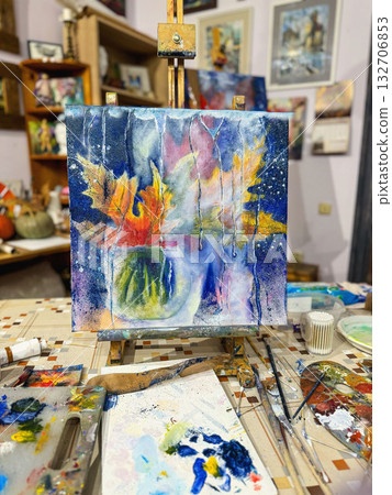 Abstract Autumn Painting on Easel in Art Studio. Colorful abstract painting with autumn leaves on easel in creative studio full of brushes, palettes. Concept of painting, fall inspiration, art freedom 132706853