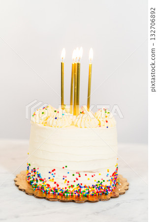 A small frosted cake sits on a white surface with tall gold candles lit at the center. The cake is decorated with piped swirls and colorful nonpareils along the base, set against a plain white wall 132706892